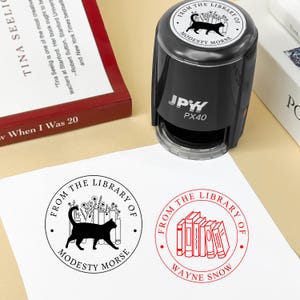May include: A black self-inking stamp with the text "FROM THE LIBRARY OF MODESTY MORSE" and a cat silhouette. The stamp is shown with two stamped impressions on white paper, one with a cat and books, and another with books.