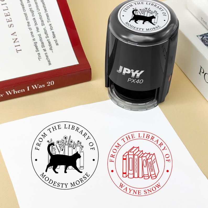 May include: A black self-inking stamp with the text "FROM THE LIBRARY OF MODESTY MORSE" and a cat silhouette. The stamp is shown with two stamped impressions on white paper, one with a cat and books, and another with books.
