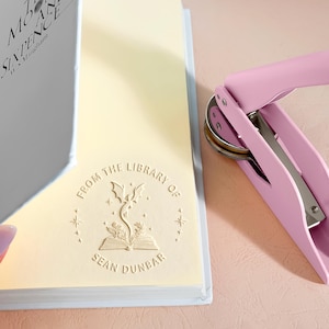 May include: A light pink book embosser next to a cream-colored book page with the text "FROM THE LIBRARY OF SEAN DUNBAR" and a dragon and book design. The book's title is "MOON AND SIXPENCE" by W.S. Maugham.