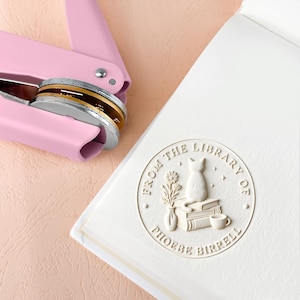 May include: A pink seal press next to a white book with a debossed design. The design includes a cat, books, flowers, and the text "FROM THE LIBRARY OF PHOEBE BIRRELL". The seal press has a silver and gold seal.