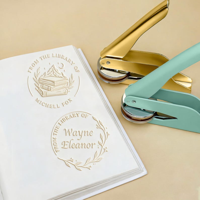 May include: A gold and a teal library seal sit next to a white book with embossed text. The book has two embossed library seals with the text "From the Library of" and names. The gold seal is open, and the teal seal is closed.