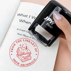 May include: A black stamp with the text "JPY PX40" is held over an open book. The book has the text "What I W When" and a red circular stamp that reads "FROM THE LIBRARY OF DREW ARCHIBALD" with a stack of books.