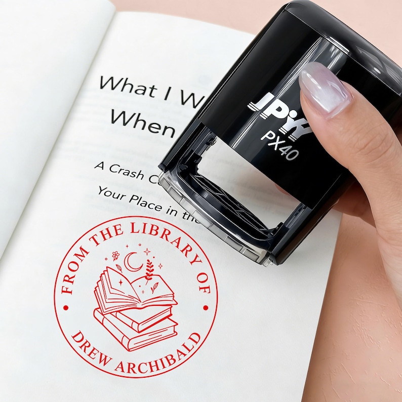 May include: A black stamp with the text "JPY PX40" is held over an open book. The book has the text "What I W When" and a red circular stamp that reads "FROM THE LIBRARY OF DREW ARCHIBALD" with a stack of books.