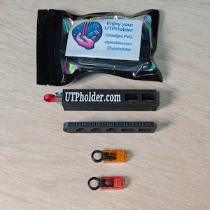 Op de afbeelding: Een assortiment zwarte elektronische componenten, waaronder een houder met de tekst "UTPholder.com", een kleiner onderdeel met de tekst "BMC NIC1 NIC2 SFP1 SFP2" en twee kleine, kleurrijke connectoren. Een verpakking met een logo en tekst is zichtbaar.