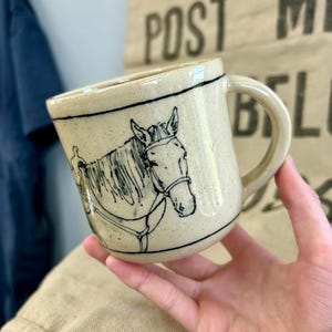 Puede incluir: Una taza de cerámica color crema moteada con un dibujo de líneas negras de un caballo con brida y silla de montar. La taza tiene un asa redondeada en el lado derecho y finas líneas negras horizontales que rodean los bordes superior e inferior.