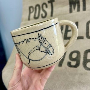 Puede incluir: Una taza de cerámica beige moteada con un dibujo de línea negra de una cabeza de caballo con brida. La taza tiene un asa redondeada y una línea negra horizontal que rodea la parte superior del exterior. La superficie tiene una textura natural.