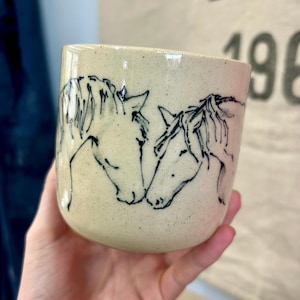 May include: A cream-colored ceramic mug with a speckled finish featuring a black line drawing of two horses facing each other. The horses are depicted in profile with detailed manes and facial features, wrapping around the front of the mug.
