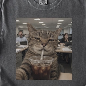 Pode incluir: T-shirt cinza escuro com uma imagem divertida de um gato bebendo café gelado com um canudo. O gato parece estar tirando uma selfie em um escritório. A camisa tem um visual vintage e desgastado.