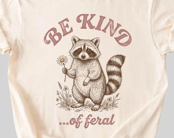 May include: Ivory-colored t-shirt featuring a raccoon holding a daisy. The text "BE KIND ...of feral" is printed above and below the raccoon illustration in a faded maroon color. The shirt has short sleeves and a crew neck.