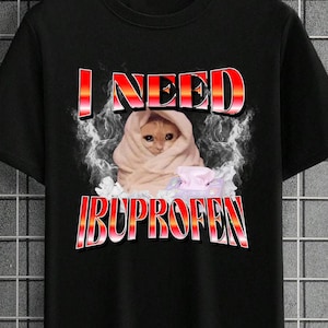 Pode incluir: Camiseta preta com o texto "I NEED IBUPROFEN" em vermelho e branco. Um gato enrolado em um cobertor está no centro do design. O design tem um efeito esfumaçado e envelhecido.