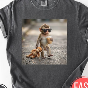 Pode incluir: Camiseta cinza escuro com uma imagem quadrada de um macaco usando óculos de sol e segurando um café gelado. Um macaco de pelúcia está no chão. A camiseta tem um visual vintage e desgastado.