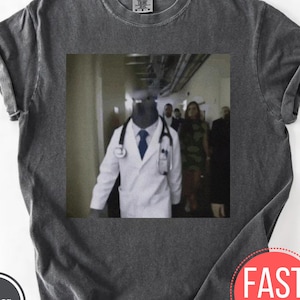 Pode incluir: T-shirt cinzento escuro com uma imagem humorística de um pombo vestido de médico, usando um jaleco branco, estetoscópio e gravata. A imagem é ambientada em um corredor com outras pessoas ao fundo. A camisa tem um visual vintage.