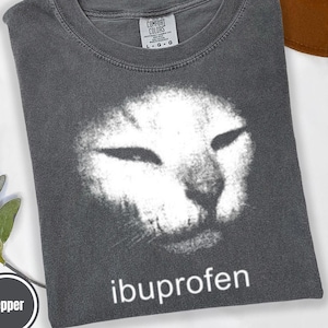 Pode incluir: Uma caneca de cerâmica branca com uma alça preta e o texto "But First, Coffee" em preto, com uma fonte manuscrita. A caneca está sobre uma superfície branca.