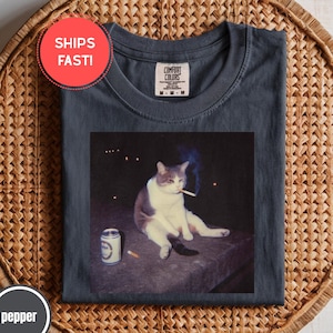 Pode incluir: Camiseta Comfort Colors cinza escuro com um gráfico de um gato fumando um cigarro, uma lata de cerveja e um cigarro em uma superfície. A camiseta tem gola redonda e uma etiqueta Comfort Colors.
