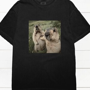 Pode incluir: T-shirt preto com um gráfico quadrado de dois marmotas num campo gramado. Um marmota está em pé, o outro parece tocá-lo. Os marmotas são castanhos e bege, o fundo é verde.