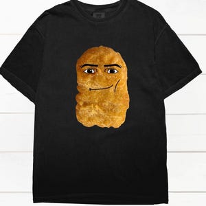 Pode incluir: Camiseta preta com um desenho de um nugget de frango sorrindo. O nugget tem uma cor marrom dourada e está centralizado na camiseta. O design é simples e divertido, adequado para uso casual. A camiseta é feita de material macio.