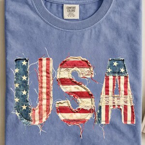 Peut inclure: T-shirt bleu Comfort Colors avec le mot "USA" appliqué sur le devant. Les lettres sont faites de denim et de tissu à rayures rouges et blanches, rappelant le drapeau américain. Le t-shirt a un col rond et une étiquette.