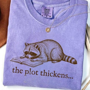 May include: A violet short-sleeve t-shirt with a crew neckline featuring a brown graphic of a raccoon reading an open book. Below the raccoon, the text reads "the plot thickens..." in brown letters. The shirt has a Comfort Colors brand label.