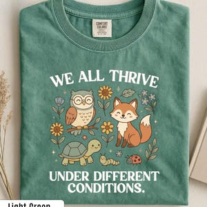 Peut inclure: Un t-shirt vert clair avec une impression graphique représentant un hibou à lunettes, un renard, une tortue, une chenille et une coccinelle entourés de fleurs. Le texte indique WE ALL THRIVE UNDER DIFFERENT CONDITIONS. Étiquette Comfort Colors.
