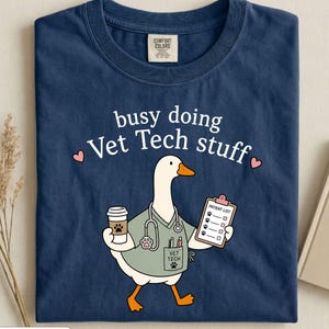 May include: A navy blue t-shirt featuring a graphic of a white goose dressed as a veterinary technician. The goose wears a light green scrub top, a stethoscope, and holds a coffee cup and a patient list clipboard. Text reads: busy doing Vet Tech stuff.