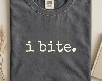 May include: A dark gray t-shirt with the text "i bite." in a white, slightly distressed font. The shirt has a crew neck and a tag that reads "Comfort Colors." The color of the shirt is "Pepper."