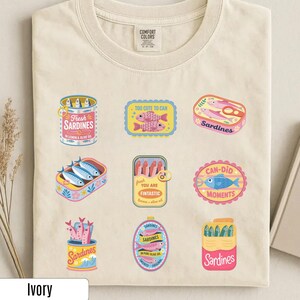 May include: Ivory t-shirt with various sardine-themed illustrations in vibrant colours. The designs include text such as "Fresh Sardines", "Too Cute to Can", and "Can-Did Moments".