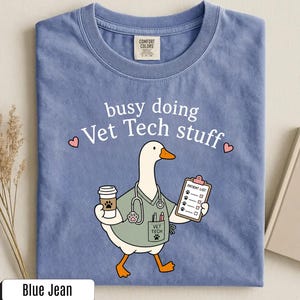 May include: A blue jean colored t-shirt featuring a graphic of a white goose dressed as a veterinary technician. The goose wears a sage green scrub top with a stethoscope, holds a coffee cup and a patient list clipboard, and has the text busy doing Vet Tech stuff printed above it.