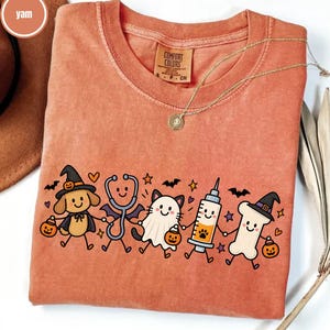 Puede incluir: Una camiseta de manga corta color ñame con un gráfico de un perro con sombrero de bruja, un estetoscopio, un gato fantasma, una jeringa y un hueso con sombrero de bruja sosteniendo calabazas. La camiseta tiene una etiqueta Comfort Colors.