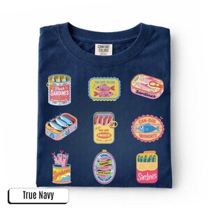 May include: A navy blue t-shirt featuring various sardine-themed designs. The graphics include sardine cans and boxes with phrases such as "Fresh Sardines" and "Too Cute to Can". The shirt is labelled "True Navy".