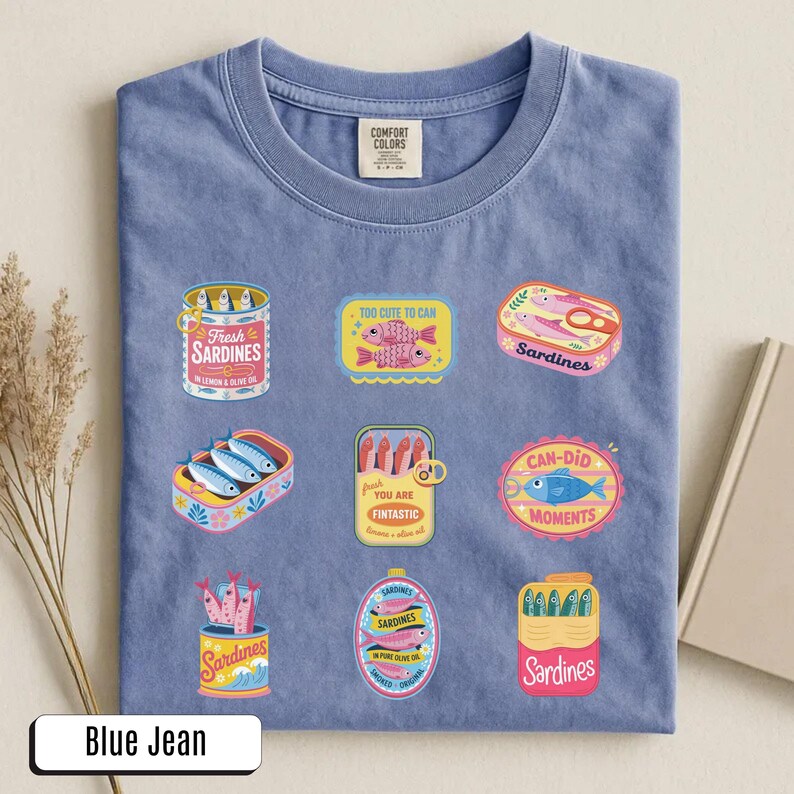 May include: Blue jean coloured t-shirt featuring various sardine-themed graphics. The graphics include sardine cans and phrases like "Too Cute to Can" and "Can-Did Moments".