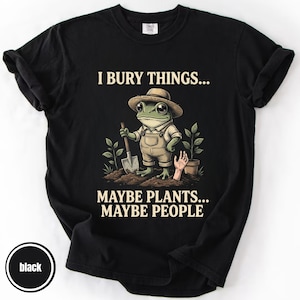 May include: Black t-shirt featuring a cartoon frog in a hat and overalls, holding a shovel. The text on the shirt says "I BURY THINGS... MAYBE PLANTS... MAYBE PEOPLE". The design also includes a hand and a potted plant.