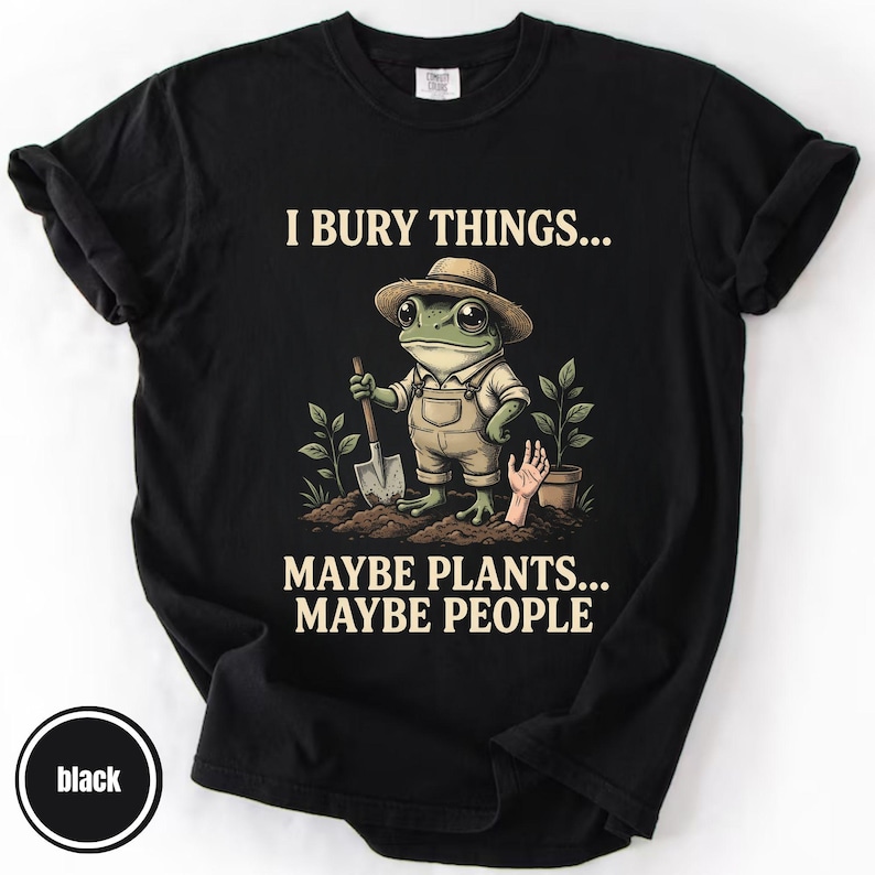 May include: Black t-shirt featuring a cartoon frog in a hat and overalls, holding a shovel. The text on the shirt says "I BURY THINGS... MAYBE PLANTS... MAYBE PEOPLE". The design also includes a hand and a potted plant.