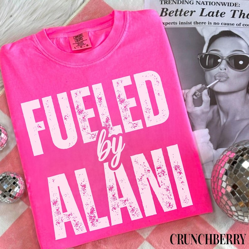 May include: A bright pink t-shirt with the words "FUELED by ALANI" in white, distressed lettering. The shirt is displayed on a pink and white checkered surface, with a newspaper clipping and a disco ball in the background. The brand name "CRUNCHBERRY" is at the bottom.