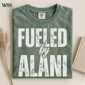 May include: A moss green t-shirt with the text "FUELED by ALANI" in white, distressed lettering. The shirt has a crew neck and a tag that reads "Comfort Colors". The shirt is folded and lying flat.