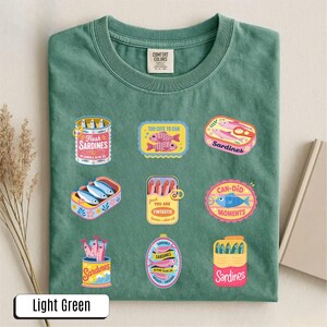 May include: Light green t-shirt with a crew neck, featuring colorful sardine can graphics. The graphics include text such as "Fresh Sardines", "Too Cute to Can", and "Can-Did Moments". The shirt is made of a soft material.