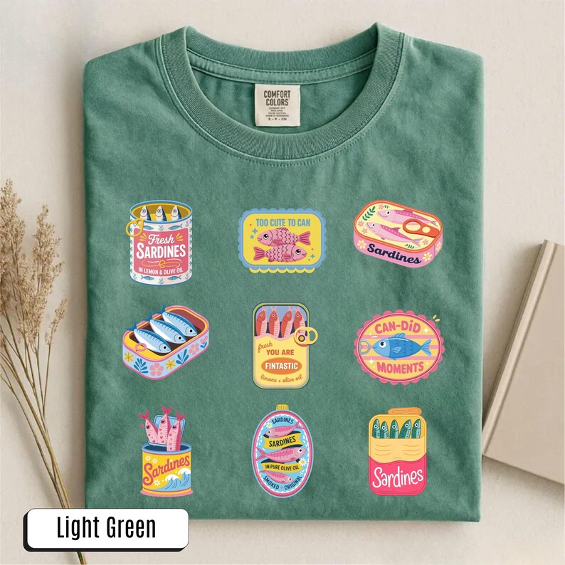 May include: Light green t-shirt with a crew neck, featuring colorful sardine can graphics. The graphics include text such as "Fresh Sardines", "Too Cute to Can", and "Can-Did Moments". The shirt is made of a soft material.