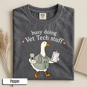May include: A charcoal gray t-shirt featuring a graphic of a white goose dressed as a veterinary technician. The goose wears a light green scrub top, a stethoscope, and holds a coffee cup and a patient list. The text reads busy doing Vet Tech stuff.