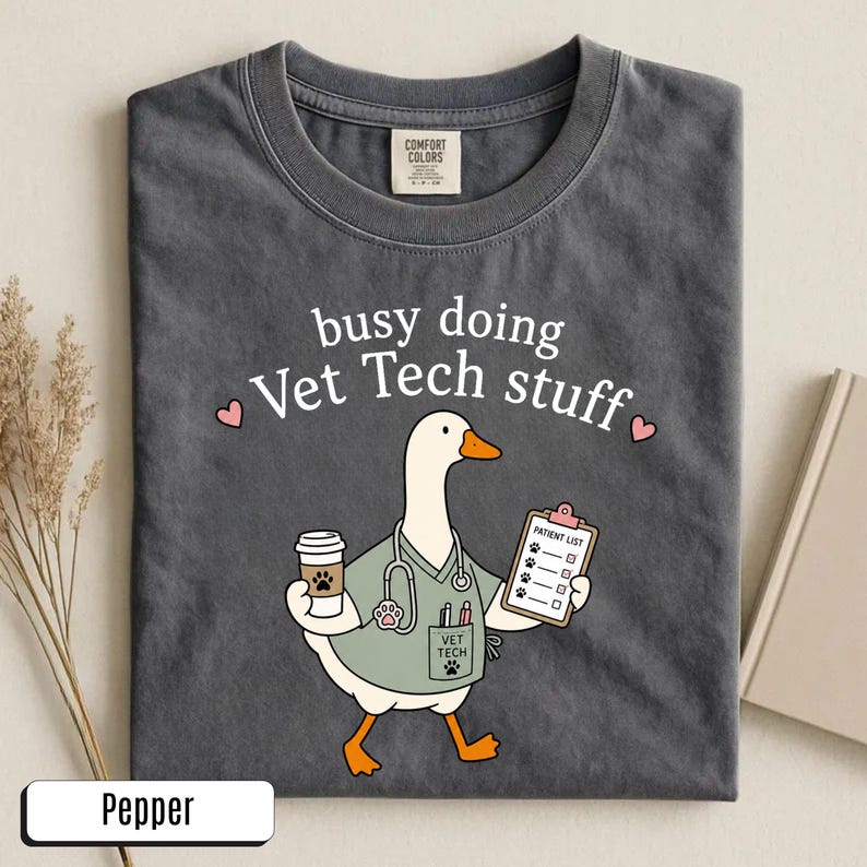 May include: A charcoal gray t-shirt featuring a graphic of a white goose dressed as a veterinary technician. The goose wears a light green scrub top, a stethoscope, and holds a coffee cup and a patient list. The text reads busy doing Vet Tech stuff.