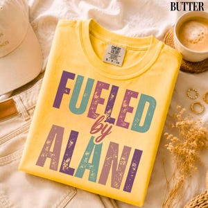 May include: A yellow t-shirt with the text "FUELED by ALANI" in teal, purple, and brown lettering. The shirt is a Comfort Colors brand, size S/P/CH. The shirt is folded on a cream-colored surface.