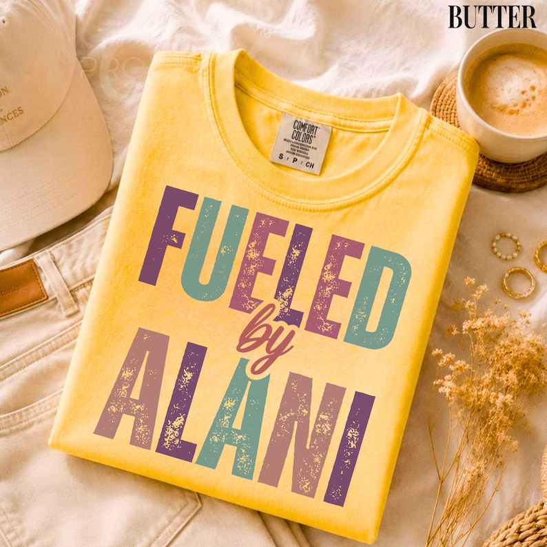 May include: A yellow t-shirt with the text "FUELED by ALANI" in teal, purple, and brown lettering. The shirt is a Comfort Colors brand, size S/P/CH. The shirt is folded on a cream-colored surface.