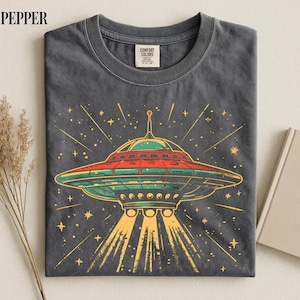 May include: A charcoal gray crew neck t-shirt featuring a graphic print of a flying saucer emitting light beams. The saucer is colored in shades of green, red, and yellow, surrounded by yellow starbursts. The inner neck label reads Comfort Colors.