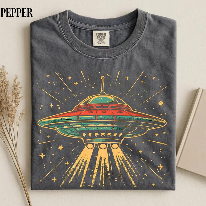 May include: A charcoal gray crew neck t-shirt featuring a graphic print of a flying saucer emitting light beams. The saucer is colored in shades of green, red, and yellow, surrounded by yellow starbursts. The inner neck label reads Comfort Colors.