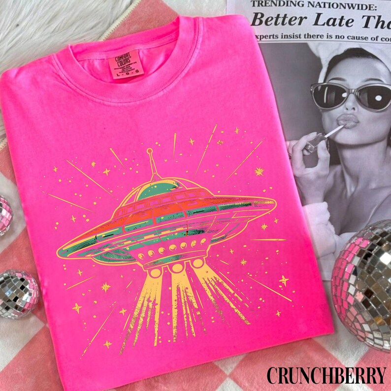 May include: A bright neon pink t-shirt featuring a graphic of a flying saucer emitting yellow light beams and surrounded by small stars. The shirt has a crew neckline and a Comfort Colors brand label printed on the inside of the collar.
