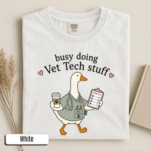 May include: A white t-shirt featuring a graphic of a white goose wearing a sage green scrub top, a stethoscope, and a pocket with a paw print. The goose holds a coffee cup and a clipboard labeled Patient List. Text reads busy doing Vet Tech stuff.