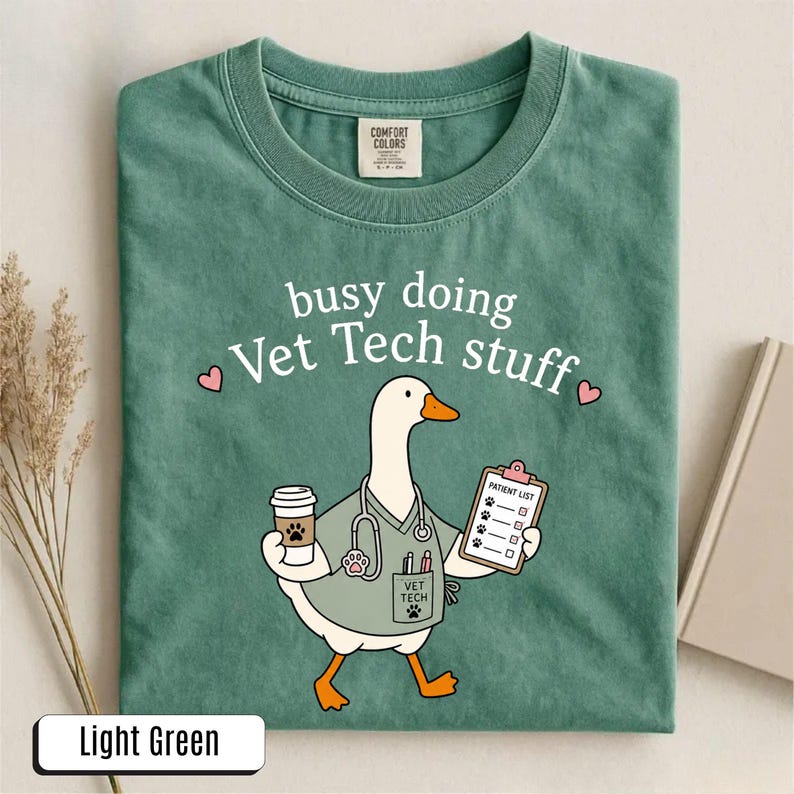 May include: A light green t-shirt featuring a graphic of a white goose dressed as a veterinary technician. The goose wears a stethoscope, holds a coffee cup and a patient list clipboard, and has the text busy doing Vet Tech stuff printed above.