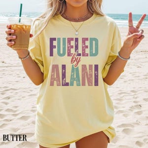 May include: Butter-colored t-shirt with the text "Fueled by Alani" in colorful block letters. The shirt is worn on a beach, and the word "BUTTER" is printed at the bottom. The shirt is a casual, oversized fit.