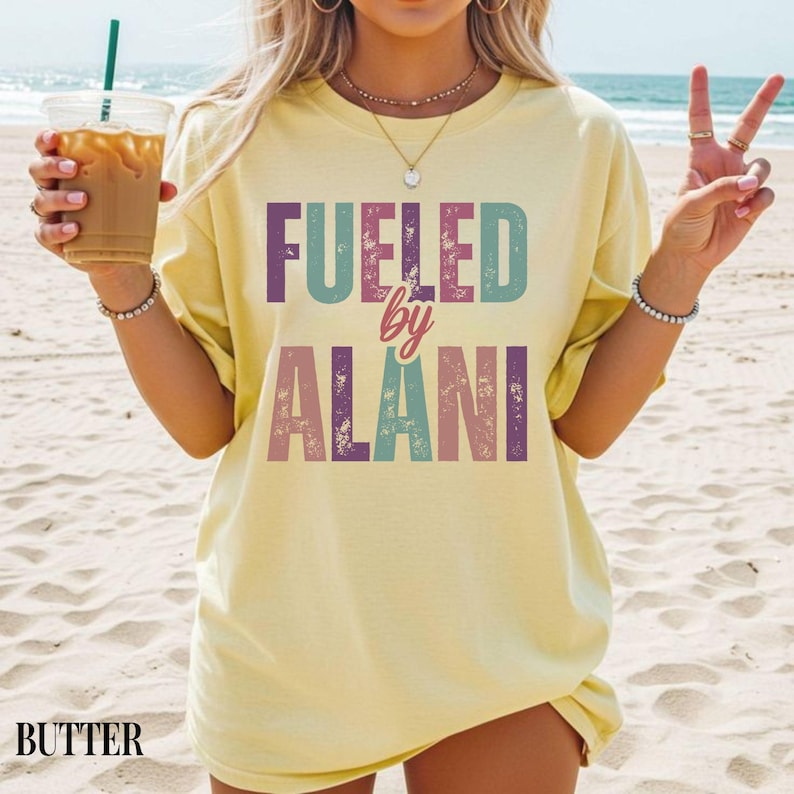 May include: Butter-colored t-shirt with the text "Fueled by Alani" in colorful block letters. The shirt is worn on a beach, and the word "BUTTER" is printed at the bottom. The shirt is a casual, oversized fit.