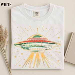 May include: A white crew neck t-shirt featuring a graphic print of a flying saucer emitting light beams. The UFO design includes red, green, and yellow colors with star accents. The shirt has a Comfort Colors brand label at the inner neckline.