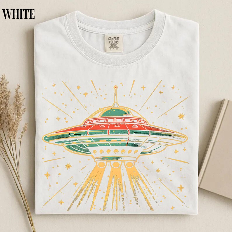 May include: A white crew neck t-shirt featuring a graphic print of a flying saucer emitting light beams. The UFO design includes red, green, and yellow colors with star accents. The shirt has a Comfort Colors brand label at the inner neckline.