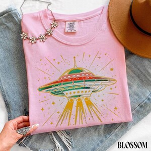 May include: A light pink t-shirt featuring a graphic of a flying saucer emitting yellow light beams and surrounded by small stars. The shirt has a crew neckline and a Comfort Colors brand label at the inner collar. The design uses red, green, and yellow.